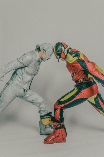 Kaksi ihmistä on vastakkain ja nojaa eteenpäin otsat vastakkain. Toisella on hopeinen futuristinen asu ja kypärä; toisella on punainen, keltainen ja musta kilpa-asu, jossa on samanlainen kypärä ja saappaat.