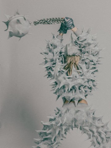 Henkilö, jolla on piikikäs, metallinhohtoinen puku ja kypärä, heiluttaa suurta piikkimailaa ketjussa yläpuolella. Tausta on tavallinen, ja poseeraus viittaa toimintaan tai liikkeeseen.