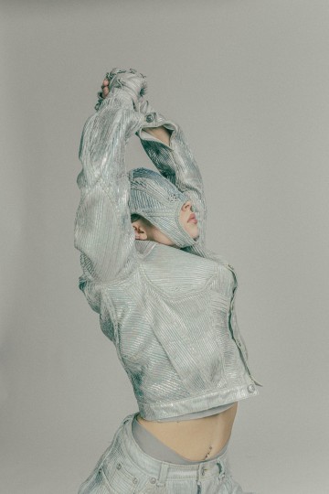 Henkilö, jolla on kuvioitu, hopeanvärinen metallipuku, poseeraa kädet ylhäällä. Hänen kasvonsa ovat suurimmaksi osaksi sopivan hupparin peitossa, ja vain nenä ja osa suusta on näkyvissä. Tausta on harmaa.