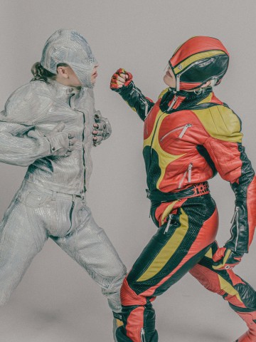 Kaksi futuristiseen panssaripukuun pukeutunutta ihmistä kohtaa toisensa. Toisella on hopeinen asu ja kypärä, kun taas toisella on punainen, keltainen ja musta puku, ja hän nostaa nyrkkiä kuin olisi lyömässä. Molemmilla on jännittynyt, dramaattinen asento.