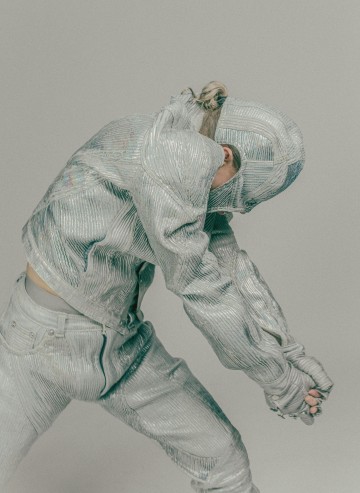 Henkilö, joka on pukeutunut kuvioituun, hopeanväriseen metalliasuun, poseeraa pää alaspäin ja kädet ojennettuina luoden dynaamisen, abstraktin asennon vaaleaa taustaa vasten. Vaatteet peittävät osittain hänen kasvonsa.