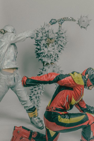 Kolme futuristiseen avantgardistiseen asuun pukeutunutta ihmistä: yksi hopeapukuinen, jolla on kädessään keihäs, yksi piikkipukuinen ja yksi punakeltainen panssaripukuinen, kaikki poseeraavat ikään kuin keskellä toimintaa vaaleaa taustaa vasten.