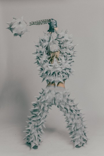 Piikkiseen, harmaaseen panssaripukuun pukeutunut henkilö heiluttaa suurta piikkimailaa. Puku peittää vartalon ja pään, ja tausta on yksinkertainen ja neutraali. Henkilön selkä on kameraan päin.