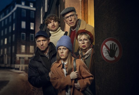 Viisi ihmistä seisoo kyyryssä hämärällä kadulla ja näyttää huolestuneelta. Heillä on talvitakit, pipot ja huivit. Heidän vieressään seinällä näkyy kyltti, jossa on käsi ja yliviivattu matkapuhelin.