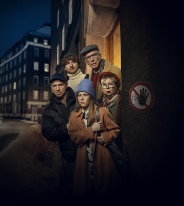 Viisi talvivaatteisiin pukeutunutta ihmistä seisoo tiiviisti vierekkäin ja kurkistelee huolestuneina yöllisen rakennuksen nurkan taakse. Seinällä on kyltti, jossa on käsi ja auton symboli. Kaupunkimainen katukuva on hämärästi valaistu.