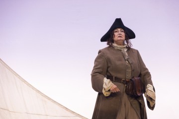 Henkilö, jolla on yllään historiallinen merirosvopuku, mukaan lukien kolmikulmainen hattu ja ruskea takki, seisoo itsevarmasti vaaleaa taustaa vasten, ja hänen takanaan näkyy osittain purje.