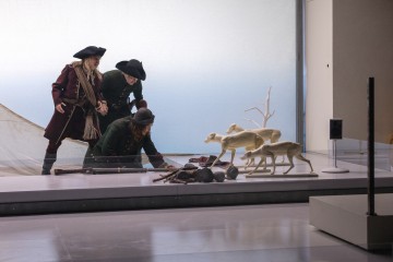 Kolme historiallisiin metsästäjiin pukeutunutta mallinukkea on asetettu poseeraamaan keinotekoisten hirvien ja rekvisiitan, kuten kiväärin ja eläinten nahkojen, viereen modernissa museonäyttelyssä.