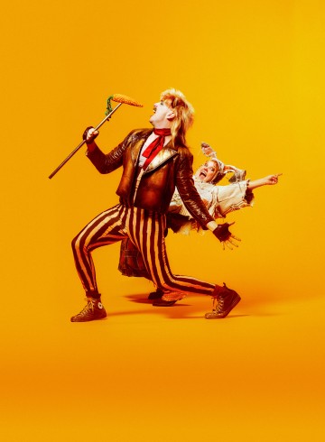 Kaksi ihmistä poseeraa dramaattisesti kirkkaan keltaista taustaa vasten. Etummaisella henkilöllä on nahkatakki ja raidalliset housut, ja hänellä on kädessään keppi, jonka päällä on maissintähkä. Takana olevalla henkilöllä on värikkäät vaatteet ja hän osoittaa ulospäin.