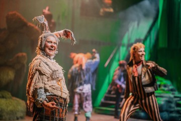 Huvittavassa narrin puvussa oleva esiintyjä hymyilee lavalla, ja hänen takanaan näkyy muita värikkäissä asuissa olevia näyttelijöitä vihreällä valaistua teatteritaustaa vasten.