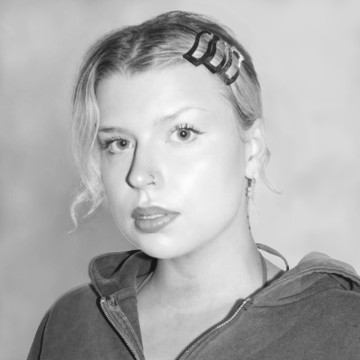 Mustavalkoinen muotokuva henkilöstä, jolla on vaaleat hiukset, jotka on muotoiltu kolmella hiusklipsillä toisella puolella, päällään vetoketjullinen huppari ja pieni korvakoru, katselee kameraan neutraalilla ilmeellä.