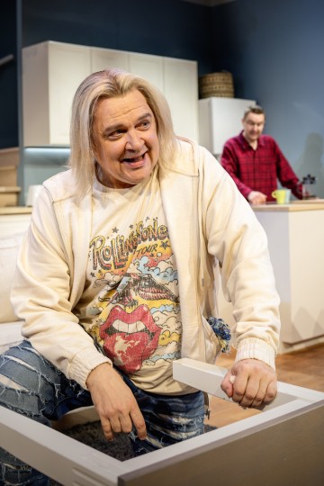 Hymyilevä henkilö, jolla on pitkät vaaleat hiukset ja Rolling Stones -paita, istuu sohvan rungolla; taustalla keittiössä seisoo toinen henkilö, jolla on punainen ruudullinen paita.