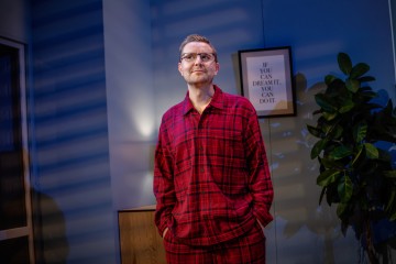 Sisätiloissa seisoo henkilö, jolla on yllään punaiset ruudulliset pyjamat. Hänen takanaan seinällä on kehystetty lainaus ja lehtikasvi. Sininen valo ja raidalliset varjot leviävät huoneen poikki.