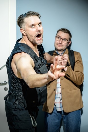 Lihaksikas mies, jolla on yllään musta taktinen liivi, kohtaa aggressiivisesti hermostuneen miehen, jolla on silmälasit ja ruskea bleiseri, tarttumalla tämän kaulukseen ja elehtimällä kädellään.
