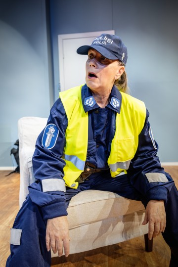 Henkilö, jolla on yllään suomalainen poliisin univormu ja keltainen liivi, istuu tuolilla ja näyttää yllättyneeltä tai huolestuneelta huoneessa, jonka seinät ovat vaaleansinisiä ja lattia puuta.