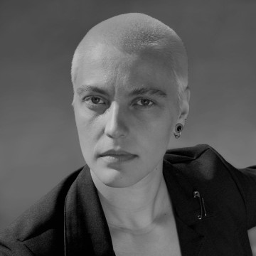 Mustavalkoinen muotokuva henkilöstä, jolla on ajeltu pää, yllään tumma bleiseri ja korvakoru, katselee hieman sivulle neutraalilla ilmeellä tavallista taustaa vasten.