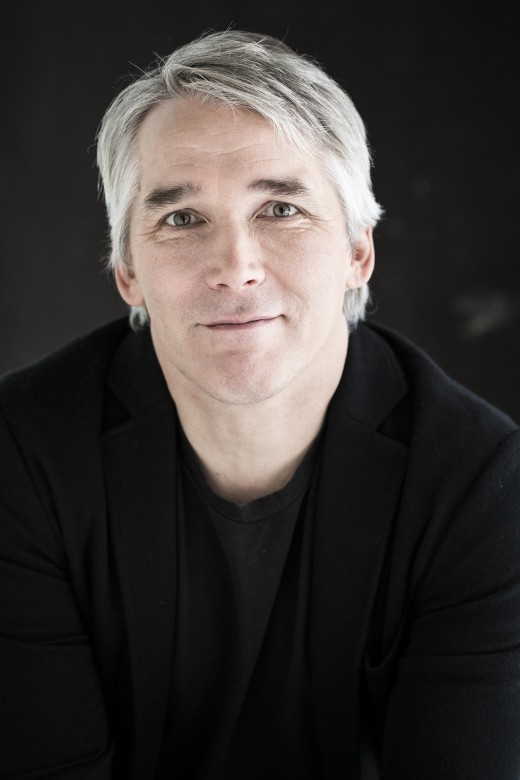 Mies, jolla on vaaleanharmaat hiukset ja vaaleat silmät, päällään musta paita ja musta takki, hymyilee lempeästi katsoessaan kameraan tummaa taustaa vasten.