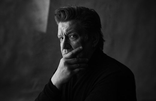 Harmaasävyinen valokuva miehestä, jolla on tyylitellyt hiukset ja vakava ilme, joka katsoo kameraan kätensä peittäessä osittain suunsa ja leukansa, tummaa, pehmeästi valaistua taustaa vasten.