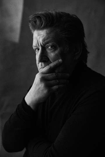 Mies, jolla on muotoiltu tukka, katsoo intensiivisesti kameraan, ja hänen kätensä peittää osittain hänen suunsa mietteliäässä asennossa. Kuva on mustavalkoinen, ja siinä on dramaattinen valaistus ja pehmeä tausta.