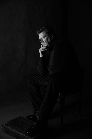Tummiin vaatteisiin pukeutunut mies istuu mietteliäänä tuolilla leuka kädellään ja ilme on mietteliäs. Kuva on mustavalkoinen ja siinä on dramaattinen valaistus ja tumma tausta.