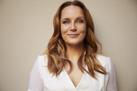 Nainen, jolla on pitkät, aaltoilevat, vaaleanruskeat hiukset, seisoo beigeä seinää vasten ja hymyilee lempeästi. Hänellä on yllään valkoinen, V-kauluksinen pusero, jossa on läpikuultavat hihat, ja hän katsoo suoraan kameraan.