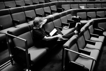 Henkilö istuu yksin tyhjässä auditoriossa, lukee tabletilta, jonka vieressä on kannettava tietokone, jalat tuolin selkänojalla, rennossa asennossa.