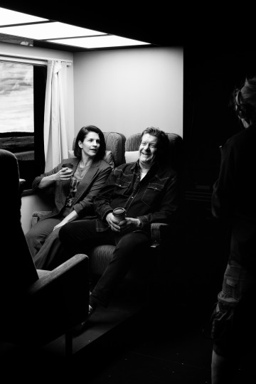 Kaksi henkilöä istuu ja keskustelee pehmeästi valaistussa huoneessa; toinen elehtii kädellään, kun taas toinen, joka pitää kädessään kuppia, hymyilee lämpimästi. Ikkuna ja toinen hahmo näkyvät osittain. Tunnelma vaikuttaa rennolta ja luonnolliselta.