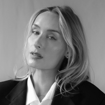 Mustavalkoinen muotokuva naisesta, jolla on vaaleat, aaltoilevat hiukset, yllään tumma bleiseri valkoisen paidan päällä, ja joka katsoo rauhallisesti kameraan tavallista taustaa vasten.