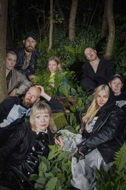 Kahdeksan ihmistä istuu ja lepää lähekkäin vihreiden kasvien ja puiden keskellä yöllisessä metsässä, pukeutuneina rentoihin ja tummiin vaatteisiin ja katsoen suoraan kameraan.
