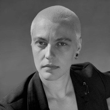 Henkilö, jolla on ajeltu pää ja vaalea iho, pukeutuu tummaan bleiseriin ja katsoo hieman sivulle. Kuva on mustavalkoinen ja siinä on neutraali, sumea tausta.