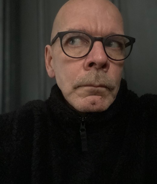 Silmälasipäinen, viiksekäs ja parranajettu henkilö pukeutuu mustaan vetoketjulliseen puseroon ja katsoo sivulle vakavalla ilmeellä. Tausta on tumma ja epätarkka.