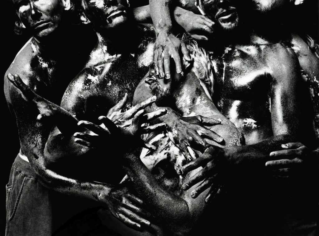 Ryhmä paidattomia ihmisiä, joiden vartalot ja kasvot on peitetty tummalla, kiiltävällä maalilla, painautuu tiiviisti yhteen ja kietoo kätensä ja käsivartensa toistensa vartaloiden yli dramaattisessa, voimakkaan kontrastisessa valaistuksessa.