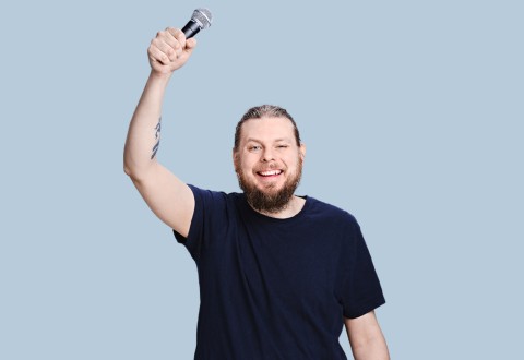 Pitkätukkainen parrakas mies, jolla on tatuointi, pitää mikrofonia toisessa kädessään, hymyilee ja iskee silmää. Hänellä on yllään tumma t-paita ja hän seisoo vaaleansinistä taustaa vasten.