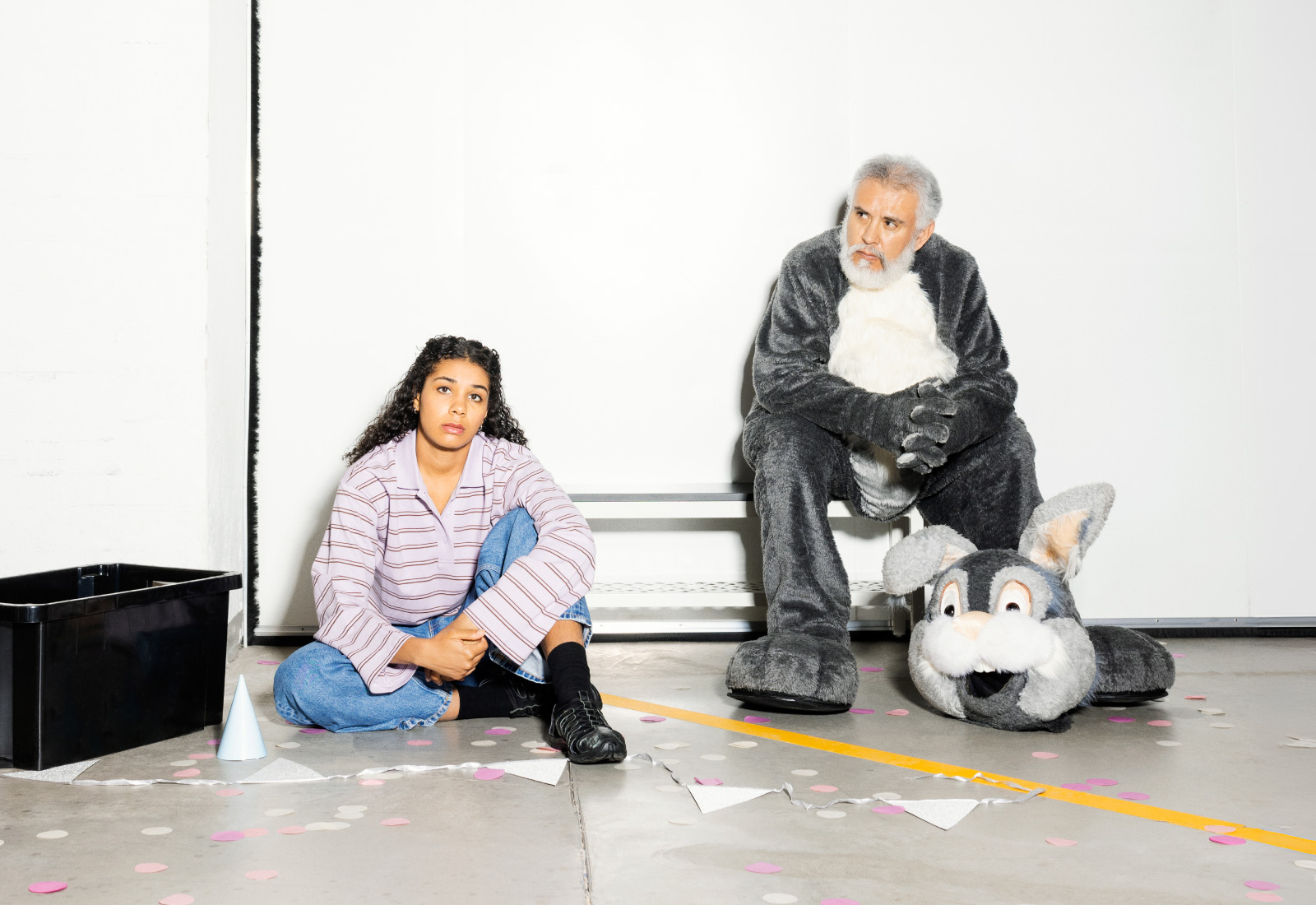 Nuori nainen istuu lattialla mietteliäänä vanhemman miehen vieressä, joka on pukeutunut kaninpukuun, josta pääosa on irrotettu; molempia ympäröivät hajallaan olevat paperikoristeet kirkkaasti valaistussa huoneessa.