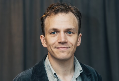 Mies, jolla on lyhyet vaaleanruskeat hiukset ja siniset silmät ja joka on pukeutunut vaaleanharmaaseen paitaan ja tummaan kuvioituun takkiin, hymyilee hieman tumman, epäselvän taustan edessä.