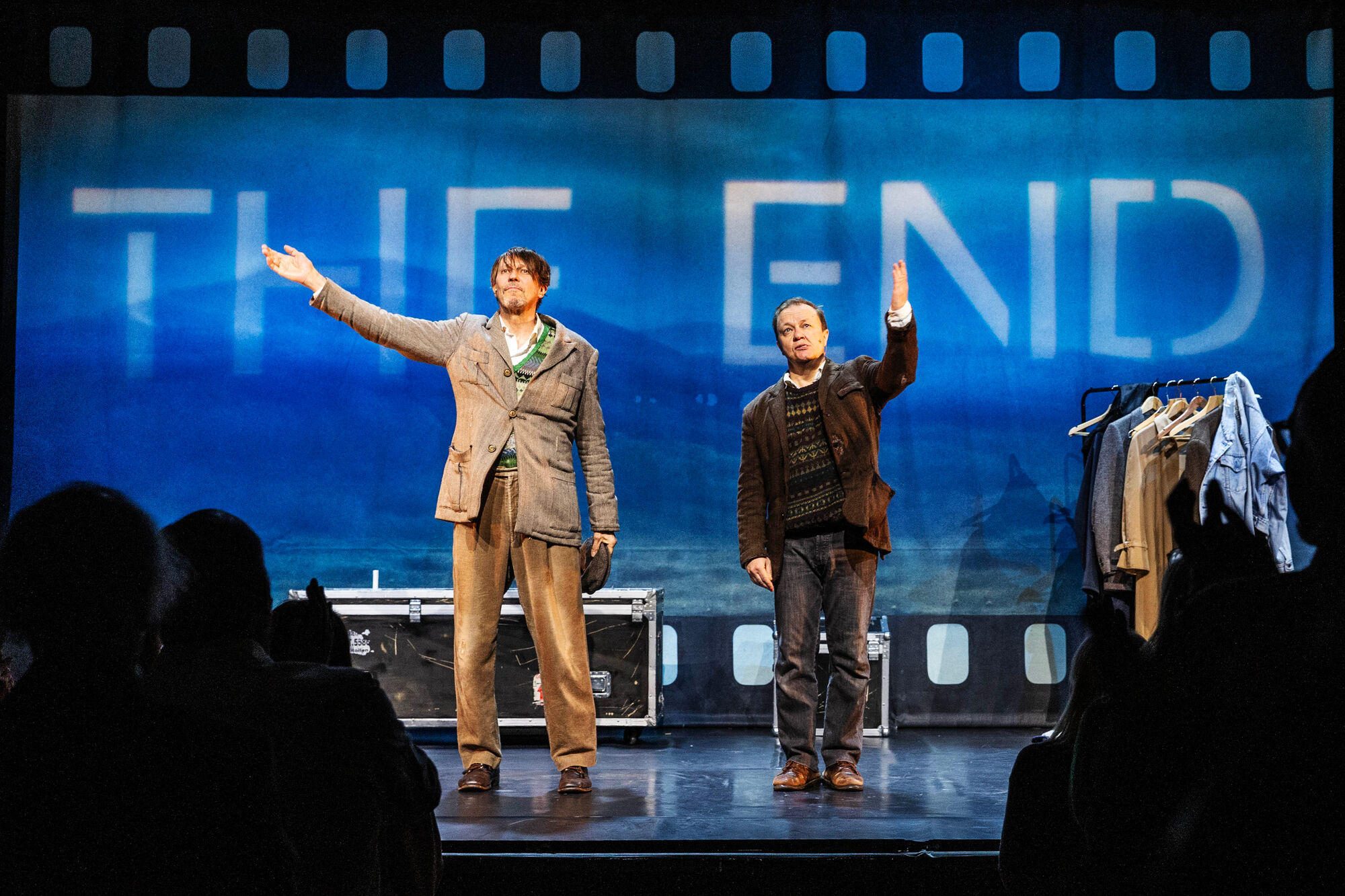 Kaksi miesnäyttelijää seisoo teatterilavalla sinisen taustakankaan edessä, jossa lukee suurilla kirjaimilla THE END.